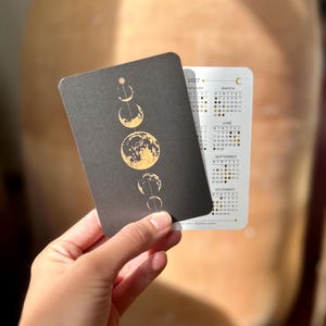 May include: A rectangular 2027 calendar card featuring a black front with a vertical gold foil illustration of moon phases. The reverse side is white with printed monthly grids for February, March, June, September, and December, including small moon icons.