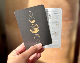 Può includere: Una carta calendario rettangolare del 2027 con un fronte nero caratterizzato da un'illustrazione verticale in lamina d'oro delle fasi lunari. Il retro è bianco con griglie mensili stampate per febbraio, marzo, giugno, settembre e dicembre, inclusi piccoli simboli lunari.