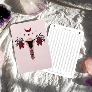 Könnte beinhalten: Eine rechteckige Karte mit hellrosa Hintergrund und einer dunkelroten anatomischen Gebärmutter-Illustration, verziert mit Blumen und Mondphasen. Daneben zeigt eine weiße 2027-Kalenderkarte Monate und Daten mit dem Text giedrestudio.com.