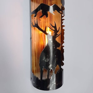Puede incluir: Una lámpara de pared rústica con un marco de metal y un panel de madera. La lámpara presenta un diseño recortado de un ciervo con cuernos y una cordillera. La lámpara está iluminada con una bombilla de luz blanca cálida.