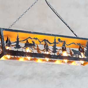 May include: A black metal chandelier with a wood top and cutouts of a forest scene, including mountains, trees, moose, and bears. The chandelier has eight light bulbs.