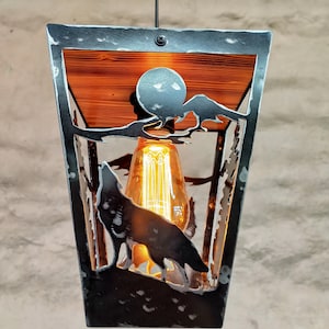 May include: A black metal pendant light with a wolf silhouette and a mountain scene. The light has a wood top and a clear glass bulb.