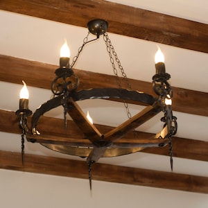 May include: A black wrought iron chandelier with a circular frame and wooden accents hangs from a wooden ceiling. The chandelier has five light bulbs and is lit.