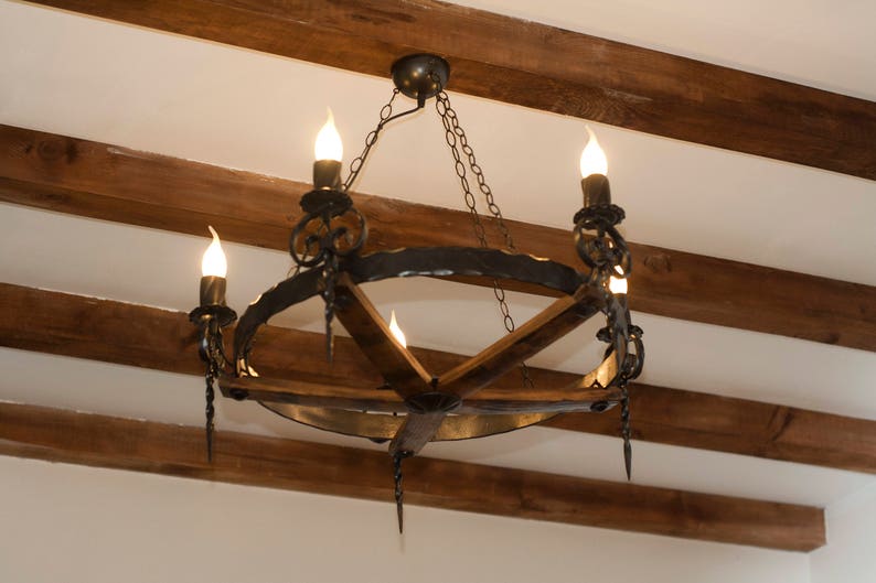 May include: A black wrought iron chandelier with a circular frame and wooden accents hangs from a wooden ceiling. The chandelier has five light bulbs and is lit.