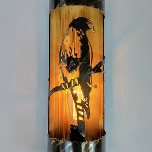 Puede incluir: Aplique de pared rústico con una silueta metálica de un halcón posado en una rama, sobre un fondo de madera. La lámpara tiene forma cilíndrica con detalles metálicos y una bombilla Edison expuesta, creando un brillo cálido.