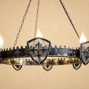 May include: A circular black metal chandelier featuring a crenelated rim and decorative shield-shaped accents with mesh centers and fleur-de-lis motifs. The fixture is suspended by chains and includes multiple flame-shaped light bulbs.