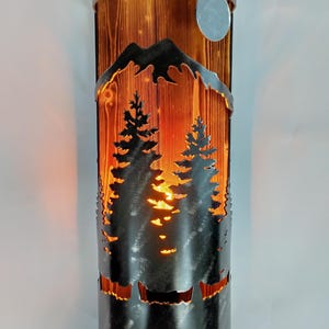 Peut inclure: Une applique murale en métal avec un fond en bois et un motif découpé d'arbres et de montagnes. L'applique a une lumière chaude qui brille à travers les découpes.