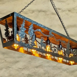 May include: Rectangular metal and wood chandelier with a forest scene cut-out design. The design includes trees, deer, and mountains. The light fixture has several exposed light bulbs and is suspended by a chain.