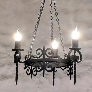 Peut inclure: Un lustre en fer forgé noir avec trois lumières en forme de bougie. Le luminaire présente des volutes décoratives et est suspendu par des chaînes. L'arrière-plan est un mur texturé et nuageux.