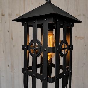 Op de afbeelding: Een matzwarte metalen hanglamp met een kooi-achtig ontwerp, voorzien van verticale staven, puntige onderranden en cirkelvormige accenten aan elke kant. De lantaarn bevat een enkele amberkleurige Edison-gloeilamp aan een ketting.