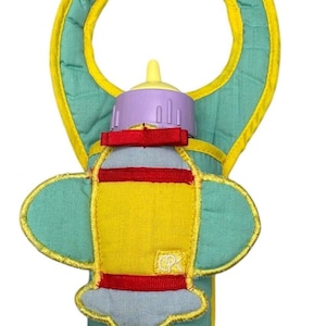 May include: A yellow and blue airplane-shaped baby bottle holder with a purple bottle inside. The holder has a red ribbon and a yellow trim. The holder has a bib attached to the top.