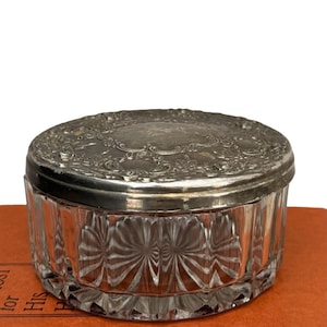 May include: A small, antique glass jar with a silver-colored lid. The clear glass jar has a ribbed design, and the lid is decorated with an ornate floral pattern. The jar is sitting on a reddish-orange surface.