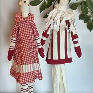 May include: Two handmade fabric dolls. One doll wears a red and white checkered dress with matching red gloves and shoes. The other doll is dressed in a red and white striped outfit with a white ruffled collar, red gloves, and shoes. Both dolls have red pointed hats.