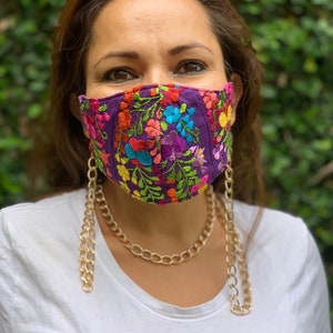 May include: A purple face mask with colorful floral embroidery, worn by a person with a gold chain necklace. The chain is attached to the mask and hangs down the front of the person's chest.