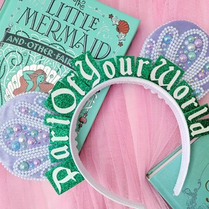 May include: A white headband with green glitter letters spelling "Part of your world". The image includes a teal book titled "The Little Mermaid" and two decorative purple felt pieces with pearl accents. The background is pink.