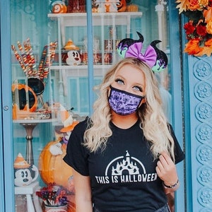 以下が含まれることがあります： ディズニーの城と「This is Halloween」という文字が白くプリントされた黒いTシャツを着た女性。彼女は、耳つきの紫と黒のヘッドバンドと、花柄の紫と黒のフェイスマスクも着用しています。女性は、ハロウィン装飾が飾られた青と白の店先に立っています。