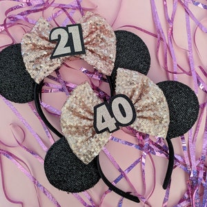 Pu&ograve; includere: Due paia di orecchie di topo nere con fiocchi di glitter rosa e oro. Un fiocco ha il numero 21 e l'altro il numero 40.