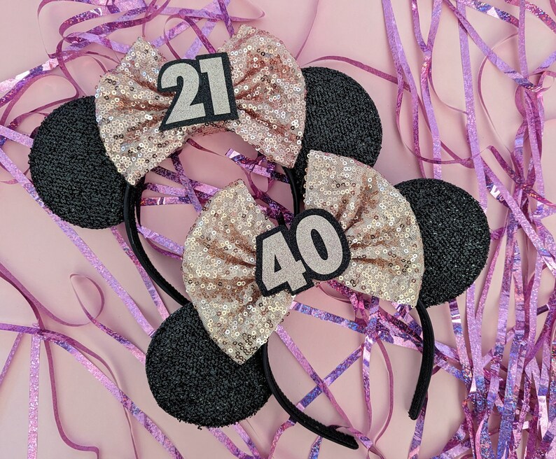 Pu&ograve; includere: Due paia di orecchie di topo nere con fiocchi di glitter rosa e oro. Un fiocco ha il numero 21 e l'altro il numero 40.