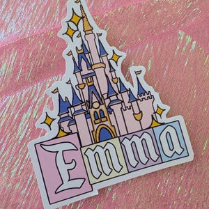 Puede incluir: Una pegatina troquelada que muestra un castillo de color rosa claro y azul con detalles dorados y formas de estrellas amarillas. Debajo del castillo, el nombre Emma est&aacute; impreso en una fuente g&oacute;tica negra, con cada letra sobre un fondo cuadrado pastel.