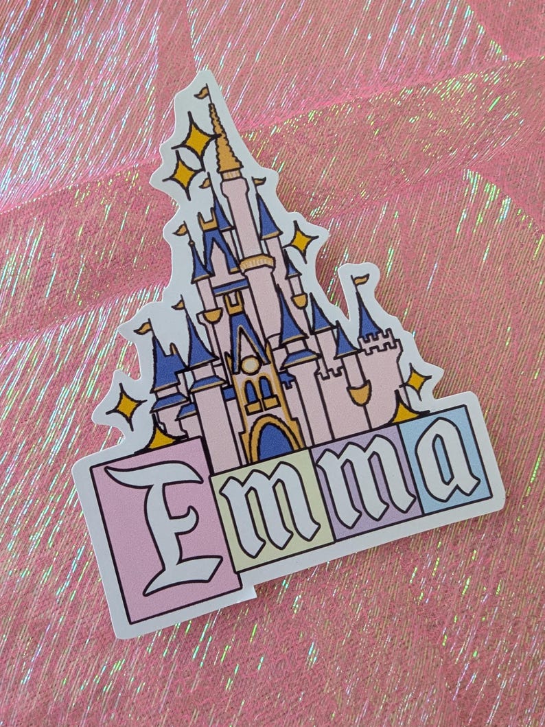 Puede incluir: Una pegatina troquelada que muestra un castillo de color rosa claro y azul con detalles dorados y formas de estrellas amarillas. Debajo del castillo, el nombre Emma est&aacute; impreso en una fuente g&oacute;tica negra, con cada letra sobre un fondo cuadrado pastel.