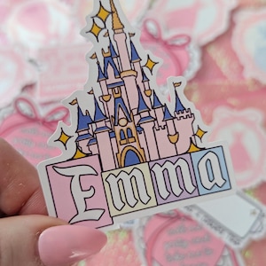 Puede incluir: Una pegatina troquelada con la ilustraci&oacute;n de un castillo rosa y azul con detalles dorados y estrellas amarillas brillantes. Debajo del castillo, el nombre Emma est&aacute; escrito en una fuente negrita con borde negro sobre bloques pastel rosa, amarillo y azul.