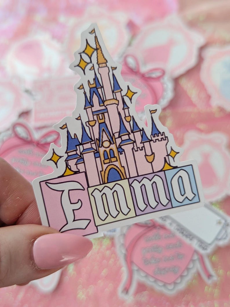 Puede incluir: Una pegatina troquelada con la ilustraci&oacute;n de un castillo rosa y azul con detalles dorados y estrellas amarillas brillantes. Debajo del castillo, el nombre Emma est&aacute; escrito en una fuente negrita con borde negro sobre bloques pastel rosa, amarillo y azul.