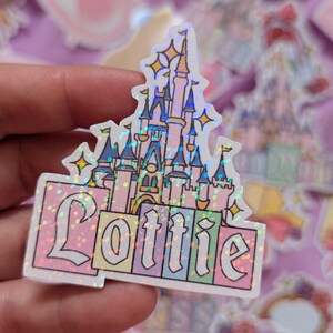 Puede incluir: Una pegatina hologr&aacute;fica con una ilustraci&oacute;n de un castillo rosa y azul con el nombre Lottie escrito en una fuente decorativa en la base. El dise&ntilde;o incluye peque&ntilde;os detalles de estrellas amarillas y un efecto de purpurina multicolor.