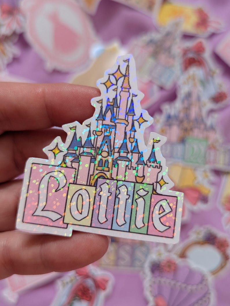 Puede incluir: Una pegatina hologr&aacute;fica con una ilustraci&oacute;n de un castillo rosa y azul con el nombre Lottie escrito en una fuente decorativa en la base. El dise&ntilde;o incluye peque&ntilde;os detalles de estrellas amarillas y un efecto de purpurina multicolor.