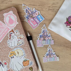 Puede incluir: Una colecci&oacute;n de pegatinas de vinilo con castillos de cuento ilustrados con los nombres Liz, Amelia y Natalie, junto a pegatinas de personajes fantasmales sobre una funda de tableta de color oro rosa brillante y una bolsa de tela blanca.