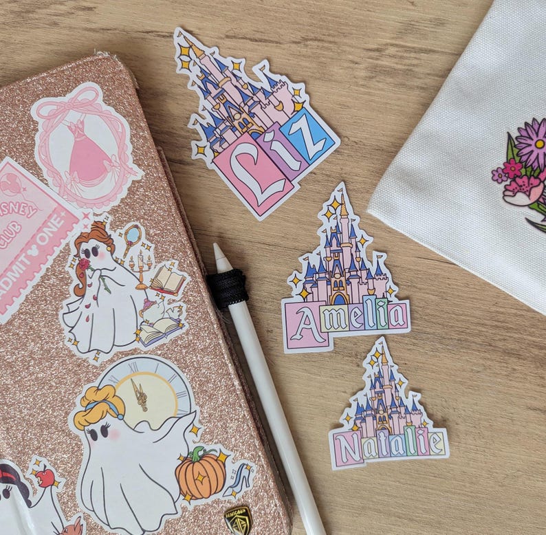 Puede incluir: Una colecci&oacute;n de pegatinas de vinilo con castillos de cuento ilustrados con los nombres Liz, Amelia y Natalie, junto a pegatinas de personajes fantasmales sobre una funda de tableta de color oro rosa brillante y una bolsa de tela blanca.