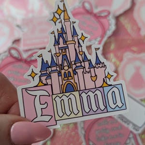 Puede incluir: Una pegatina troquelada que muestra una ilustraci&oacute;n de un castillo de cuento de hadas en rosa y azul con estrellas amarillas. Debajo del castillo, el nombre Emma est&aacute; impreso en letras negras sobre cuatro bloques cuadrados de colores pastel: rosa, amarillo, azul claro y blanco.