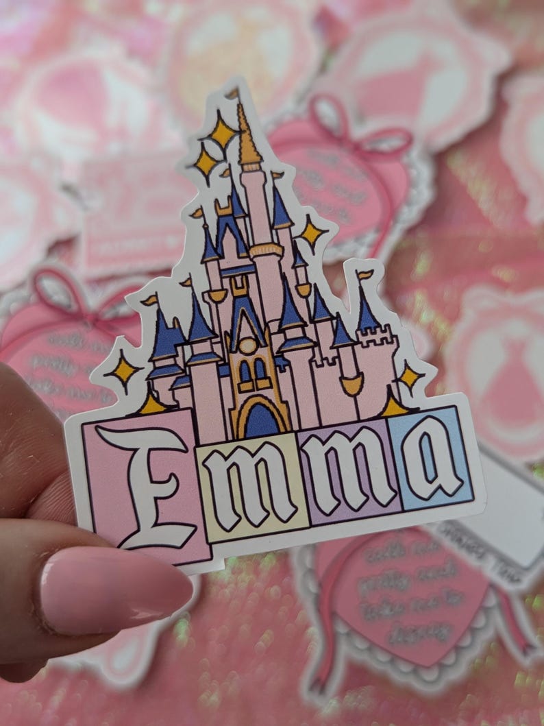 Puede incluir: Una pegatina troquelada que muestra una ilustraci&oacute;n de un castillo de cuento de hadas en rosa y azul con estrellas amarillas. Debajo del castillo, el nombre Emma est&aacute; impreso en letras negras sobre cuatro bloques cuadrados de colores pastel: rosa, amarillo, azul claro y blanco.