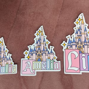 Puede incluir: Tres pegatinas troqueladas con una ilustraci&oacute;n de un castillo rosa y azul con estrellas amarillas. Cada pegatina muestra un nombre en fuente de bloque: Natalie, Amelia y Liz. Los dise&ntilde;os tienen un borde blanco y bloques de color pastel.