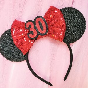 Pu&ograve; includere: Un cerchietto nero in feltro con orecchie di topo in feltro nero, decorato con un fiocco rosso di paillettes e un numero 30 in feltro nero.