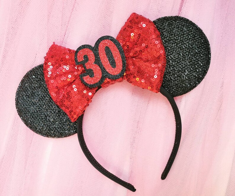 Pu&ograve; includere: Un cerchietto nero in feltro con orecchie di topo in feltro nero, decorato con un fiocco rosso di paillettes e un numero 30 in feltro nero.