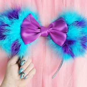 May include: A purple satin bow headband with blue and purple feather ears. The headband is on a pink tulle background.