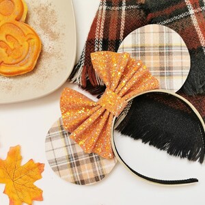 May include: An orange glitter bow headband with plaid ears, a Mickey Mouse waffle, and autumn leaves. The headband has a black base and a gold trim. A plaid scarf is in the background.