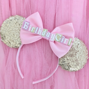 May include: A headband featuring two round ears covered in gold sequins and a large, central light pink satin bow. The bow is decorated with glittery pink and gold lettering that spells out the words Birthday Girl in a stylised font.