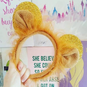 Può includere: Un cerchietto marrone con orecchie pelose e dettagli di glitter dorati. Il cerchietto è progettato per assomigliare a un leone.