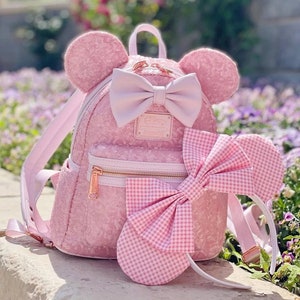 Könnte beinhalten: Rosa paillettenbesetzter Minnie Mouse Rucksack mit einer großen rosa Schleife und einer kleineren rosa-weißen Karoschleife. Der Rucksack hat einen goldenen Reißverschluss und den Schriftzug "Disney" und "Loungefly" auf der Vorderseite.
