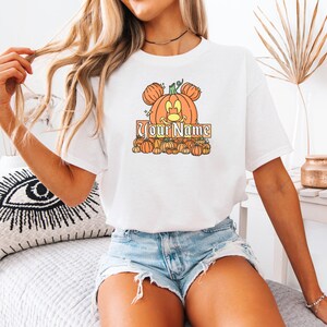 May include: White t-shirt featuring a pumpkin design with Mickey Mouse ears and the text "Your Name" in a decorative font. The design includes several small pumpkins. The shirt is a casual, comfortable style.