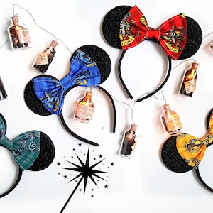 Könnte beinhalten: Vier Paar schwarze Mäuseohren mit verschiedenen farbigen Schleifen und Harry-Potter-Hauswappen. Die Schleifen sind rot, blau, grün und gelb. Die Wappen sind Gryffindor, Ravenclaw, Slytherin und Hufflepuff. Die Ohren sind mit kleinen Glühbirnen verziert.