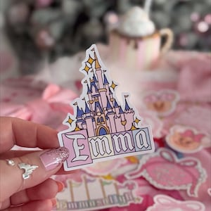 Puede incluir: Una pegatina troquelada que muestra una ilustraci&oacute;n de un castillo rosa y azul con detalles de estrellas amarillas. Debajo del castillo, el nombre Emma est&aacute; impreso en una fuente negrita y estilizada dentro de un borde rectangular blanco.