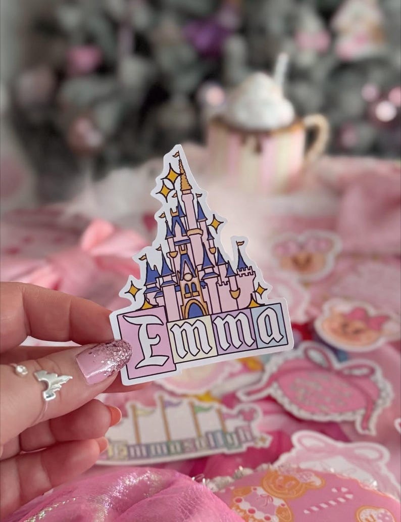 Puede incluir: Una pegatina troquelada que muestra una ilustraci&oacute;n de un castillo rosa y azul con detalles de estrellas amarillas. Debajo del castillo, el nombre Emma est&aacute; impreso en una fuente negrita y estilizada dentro de un borde rectangular blanco.