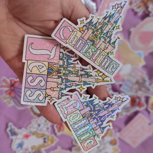 Puede incluir: Tres pegatinas hologr&aacute;ficas con un dise&ntilde;o de castillo estilizado en tonos pastel rosa, azul y amarillo. Cada pegatina muestra un nombre &uacute;nico en letras blancas: Jess, Christine y Lottie. Tienen un acabado brillante y reluciente.