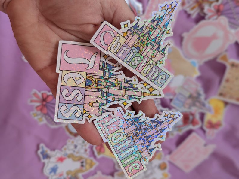 Puede incluir: Tres pegatinas hologr&aacute;ficas con un dise&ntilde;o de castillo estilizado en tonos pastel rosa, azul y amarillo. Cada pegatina muestra un nombre &uacute;nico en letras blancas: Jess, Christine y Lottie. Tienen un acabado brillante y reluciente.