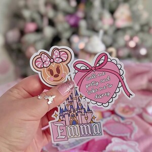 Puede incluir: Tres pegatinas troqueladas que presentan una cabeza de Minnie Mouse estilo pan de jengibre, un coraz&oacute;n rosa con un lazo y el texto "call me pretty and take me to disney", y un castillo rosa con el nombre "Emma" impreso en una fuente estilizada.