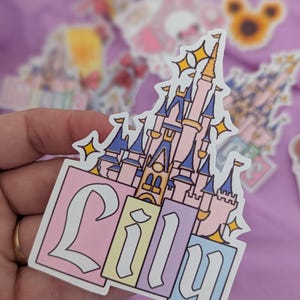 Puede incluir: Una pegatina troquelada que muestra un castillo rosa y azul con detalles dorados y estrellas amarillas brillantes. El nombre Lily est&aacute; escrito en una fuente blanca estilizada y llamativa sobre tres bloques rectangulares en rosa, amarillo y azul.
