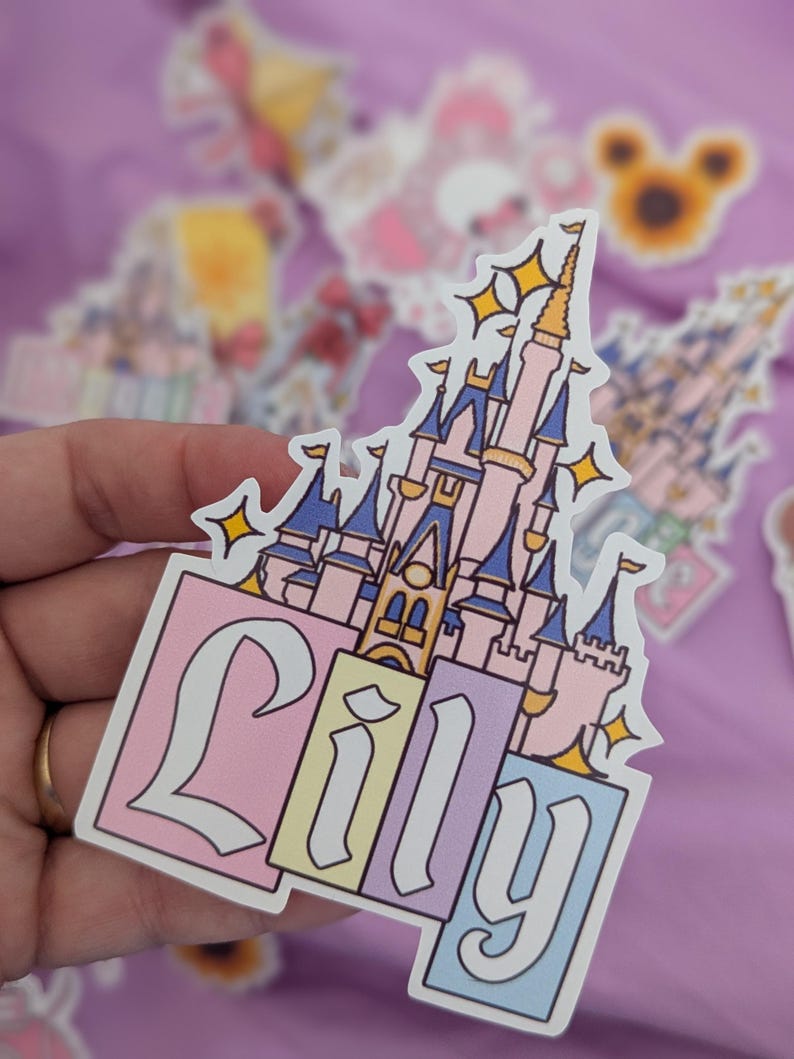 Puede incluir: Una pegatina troquelada que muestra un castillo rosa y azul con detalles dorados y estrellas amarillas brillantes. El nombre Lily est&aacute; escrito en una fuente blanca estilizada y llamativa sobre tres bloques rectangulares en rosa, amarillo y azul.