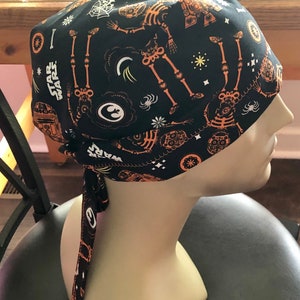 May include: Black surgical cap with a repeating orange skeleton and Star Wars themed print. The cap features a tie-back design and is displayed on a mannequin head.